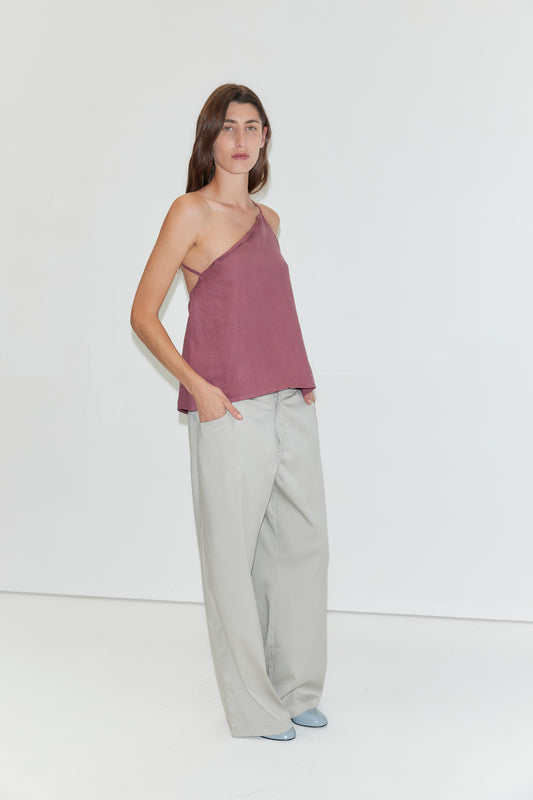 A woman stands against a white background, wearing the "Half Halter - Brick" one-shoulder top by Deiji Studios, loose light gray pants, and light blue shoes. She has long brown hair and her hands in her pockets.