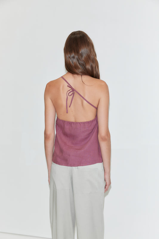 A woman stands facing away, wearing the Half Halter top in Brick by Deiji Studios and light gray pants. Her long brown hair is down, and the background is plain white.