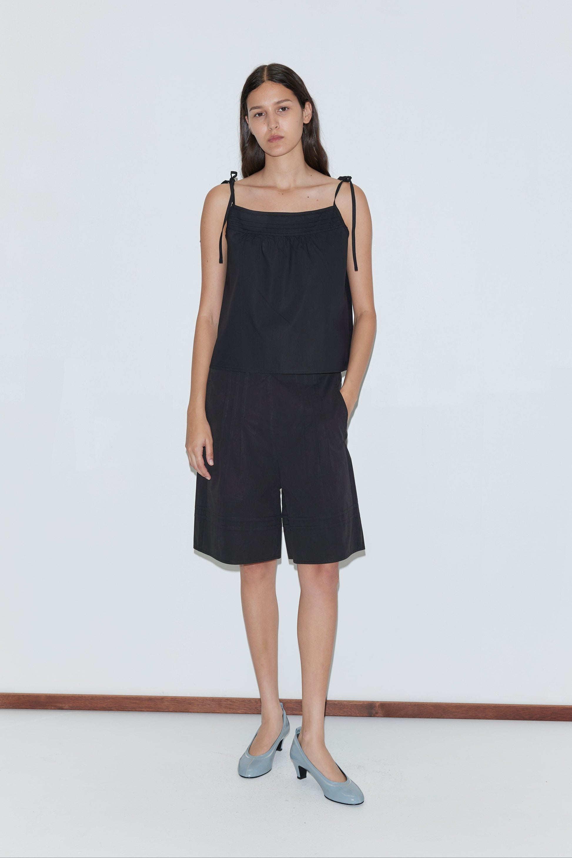 A woman with long dark hair stands against a white wall wearing a black sleeveless top, Deiji Studios Pintuck Short - Black, and light blue heels. She poses with one hand in her pocket and maintains a neutral expression.