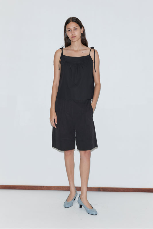 A woman with long dark hair stands against a white wall wearing a black sleeveless top, Deiji Studios Pintuck Short - Black, and light blue heels. She poses with one hand in her pocket and maintains a neutral expression.