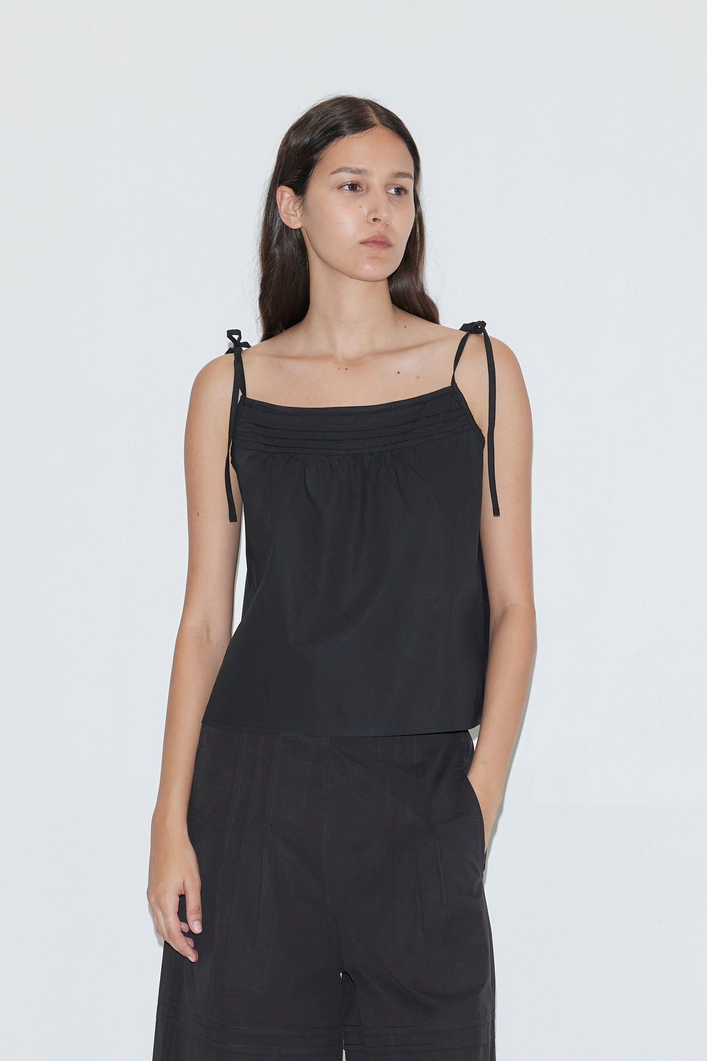 A woman with long dark hair wears the Tied Cami - Black from Deiji Studios, styled with loose black pants, and stands against a plain white background.