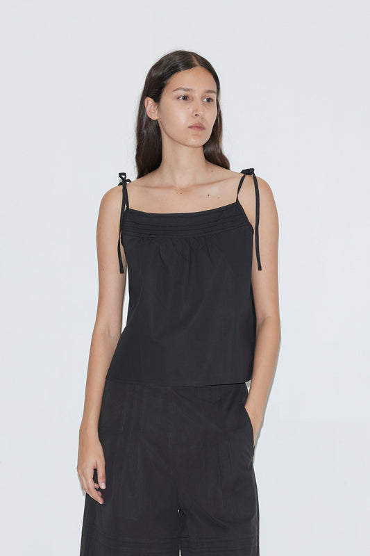 A woman with long dark hair wears the Tied Cami - Black from Deiji Studios, styled with loose black pants, and stands against a plain white background.