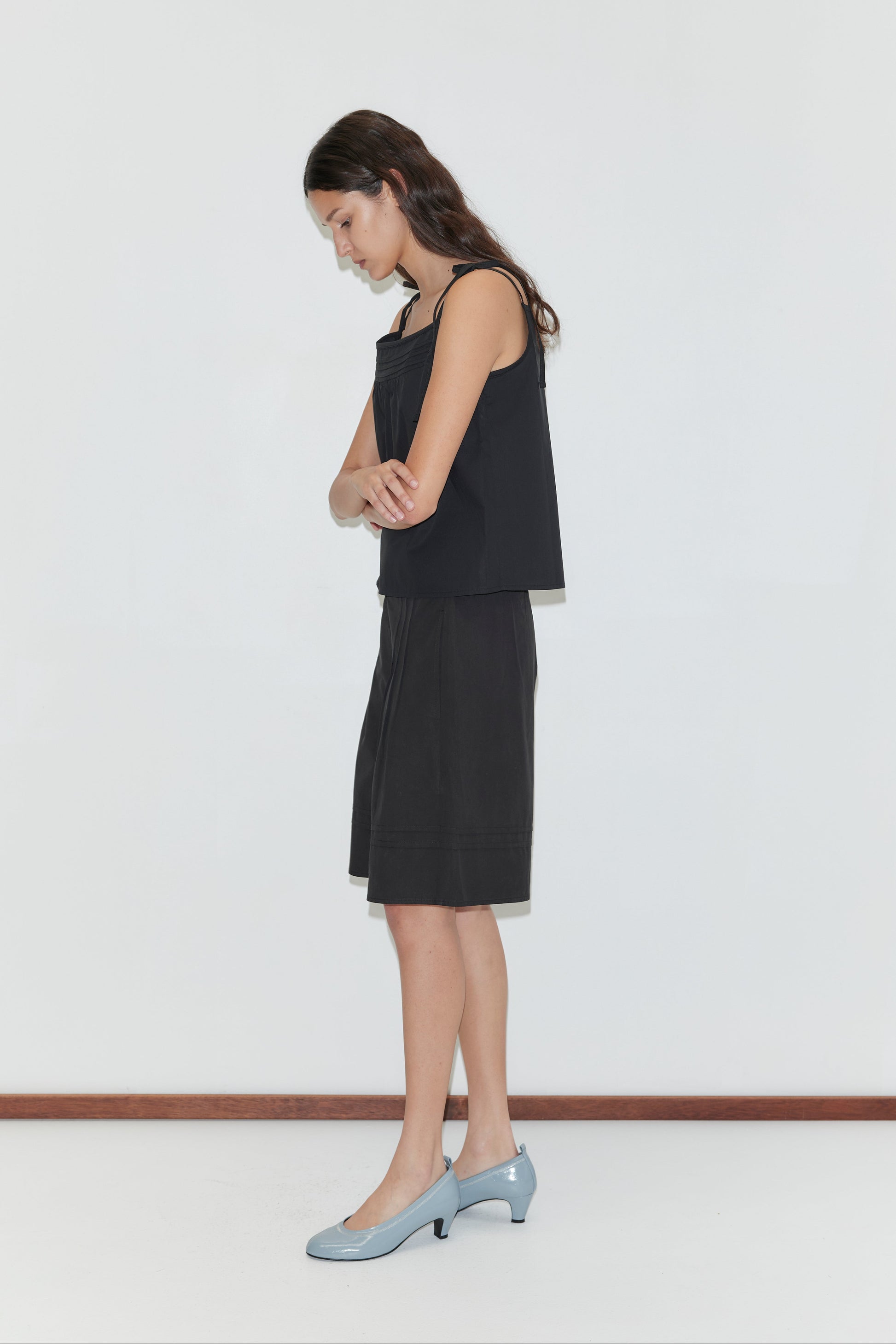 A woman with long brown hair stands in profile against a white background, arms crossed and looking down, wearing the Pintuck Short - Black. Her sleeveless top and light blue heels evoke Deiji Studios’ refined aesthetic.