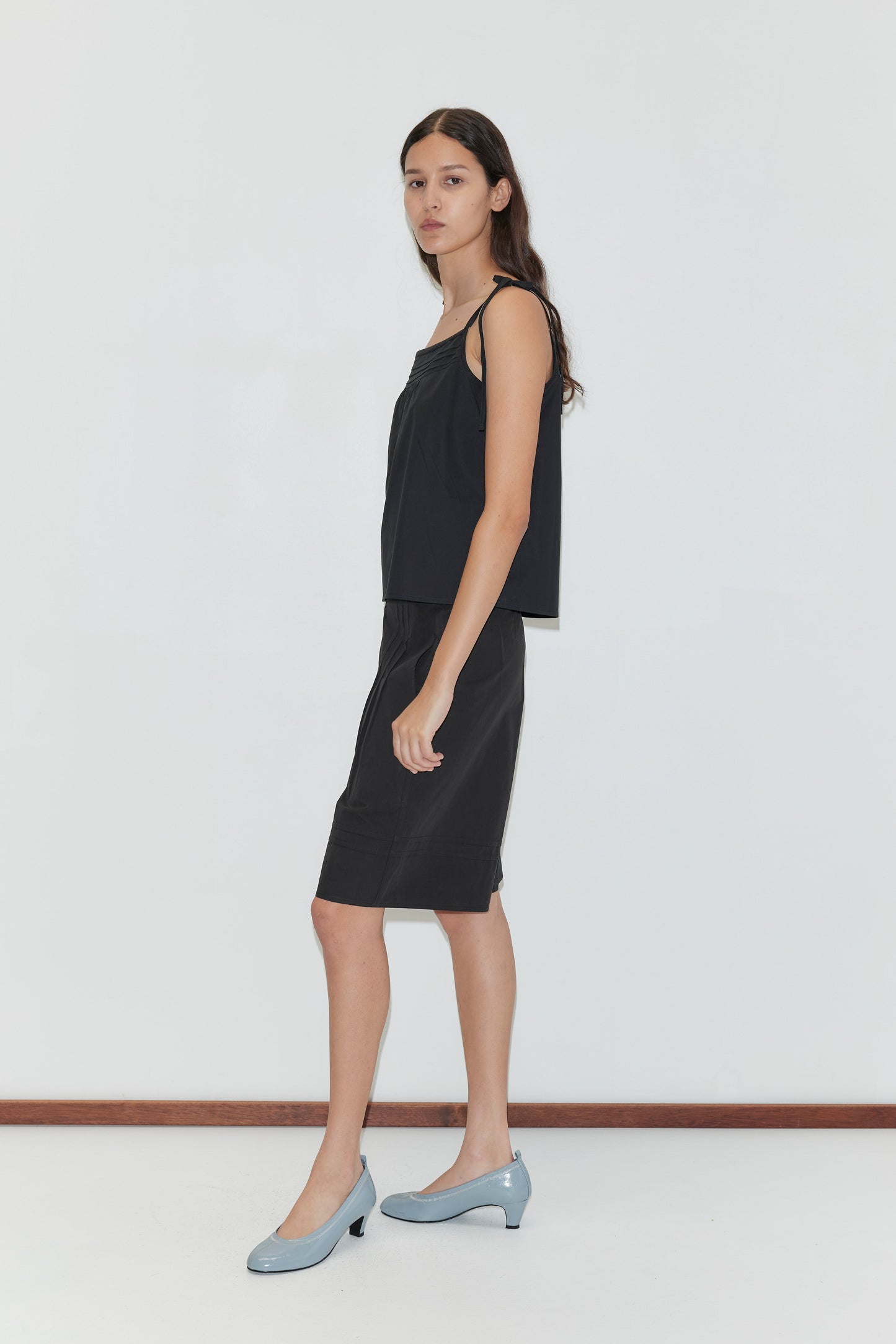 A woman with long dark hair stands sideways against a plain white wall, wearing the Tied Cami - Black from Deiji Studios, a knee-length skirt, and light blue low-heeled shoes.