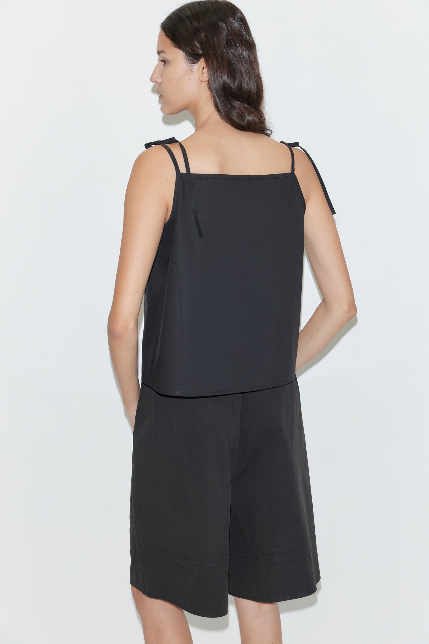 A woman with long dark hair stands facing away, wearing Deiji Studios' Tied Cami - Black with thin straps and matching black knee-length shorts against a plain white background.