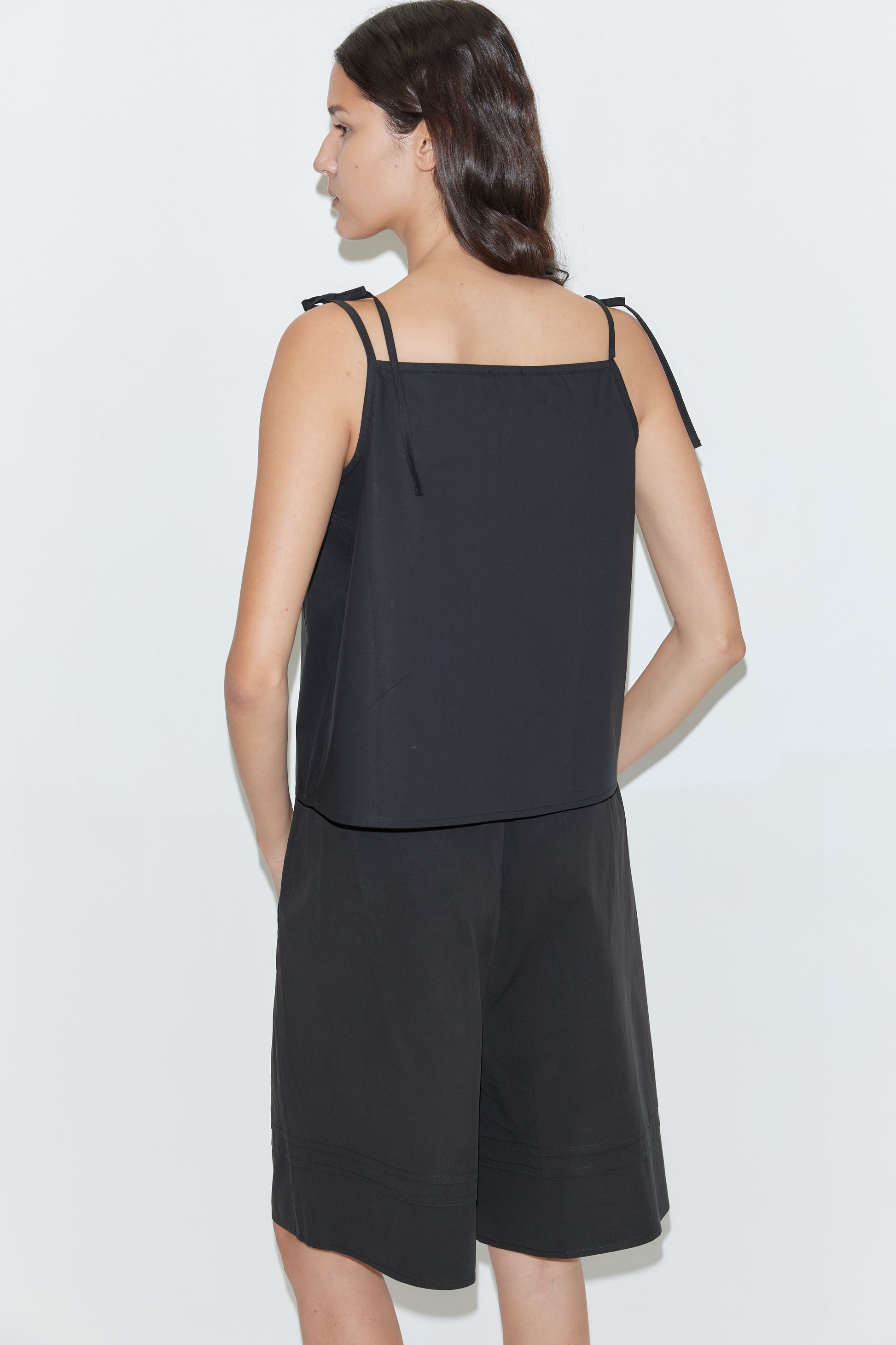 A woman with long dark hair stands facing away, wearing Deiji Studios' Tied Cami - Black with thin straps and matching black knee-length shorts against a plain white background.