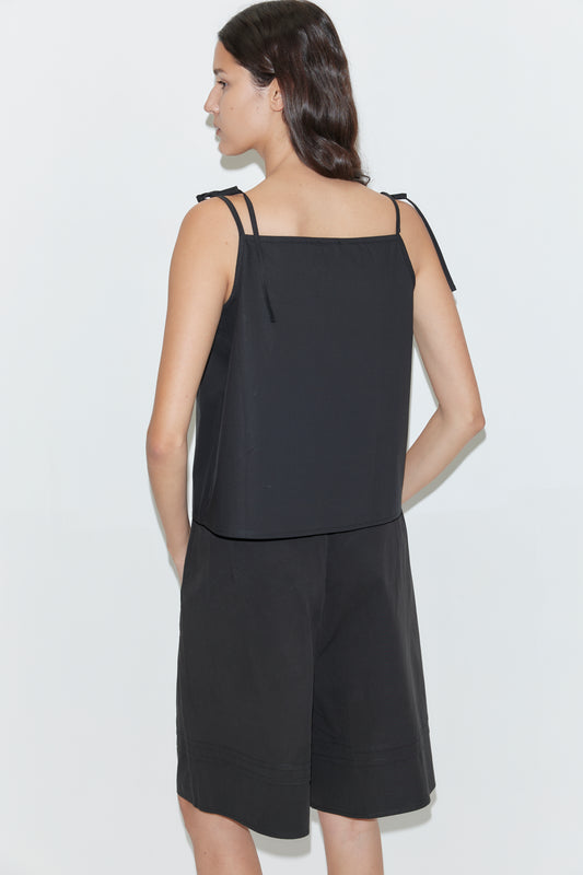 A woman with long dark hair stands facing away, wearing Deiji Studios' Tied Cami - Black with thin straps and matching black knee-length shorts against a plain white background.