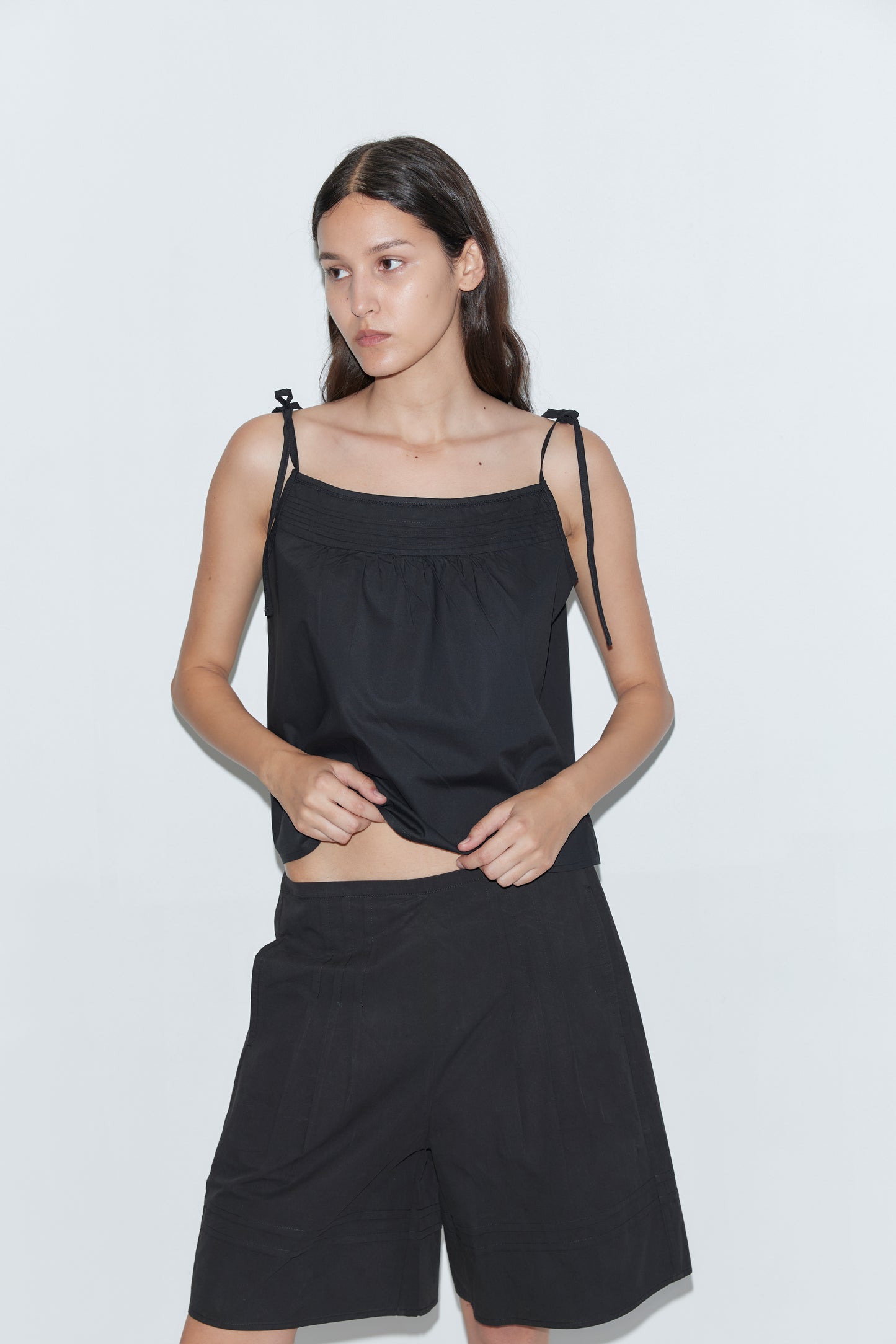 A woman stands against a plain white background wearing the Deiji Studios Pintuck Short in black and a sleeveless black top, looking to the side with her hands on her hips.