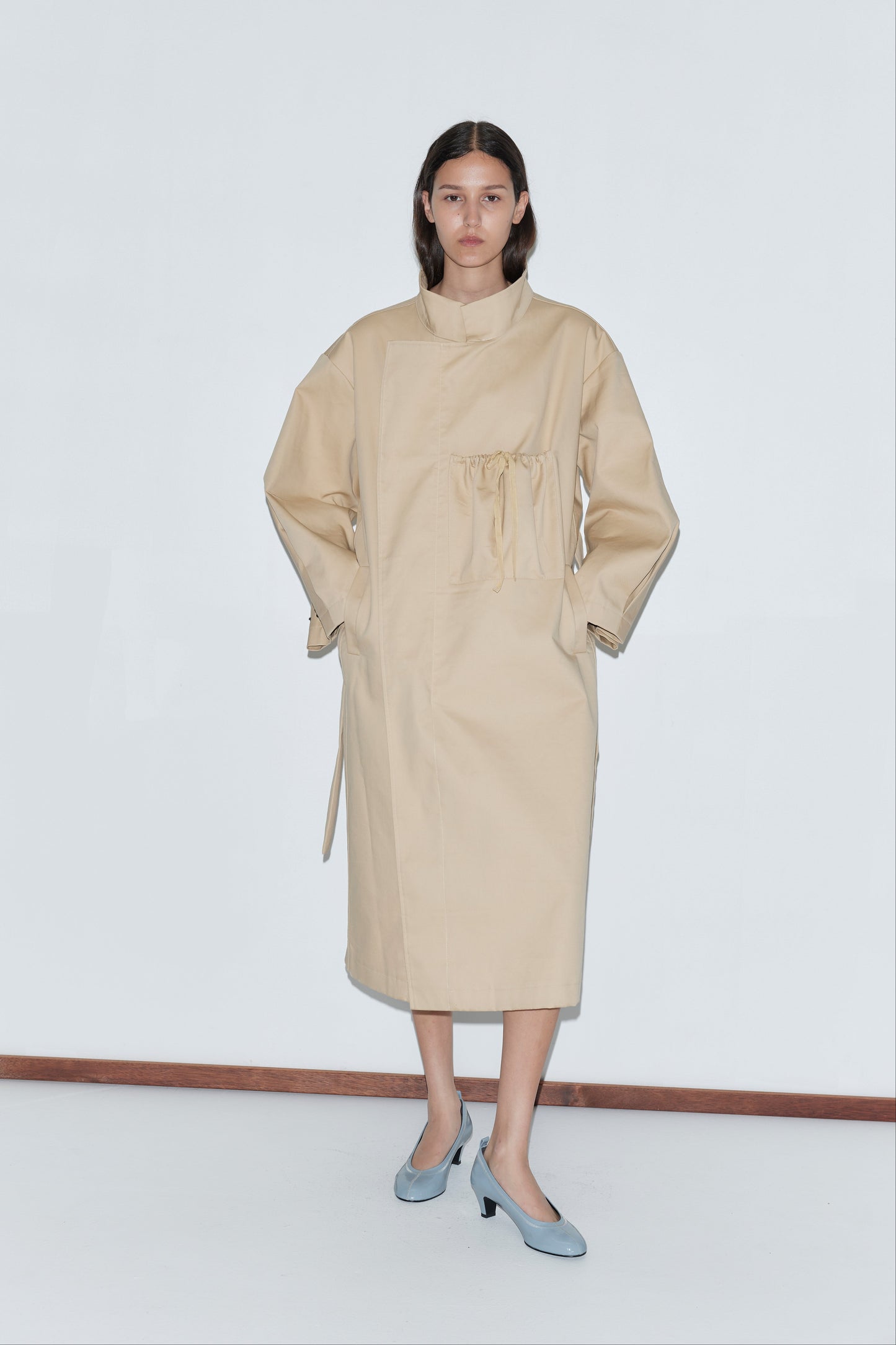 A woman stands against a white wall, wearing the Longline Jacket - Beige with a high collar and gathered waist, paired with light blue heels.