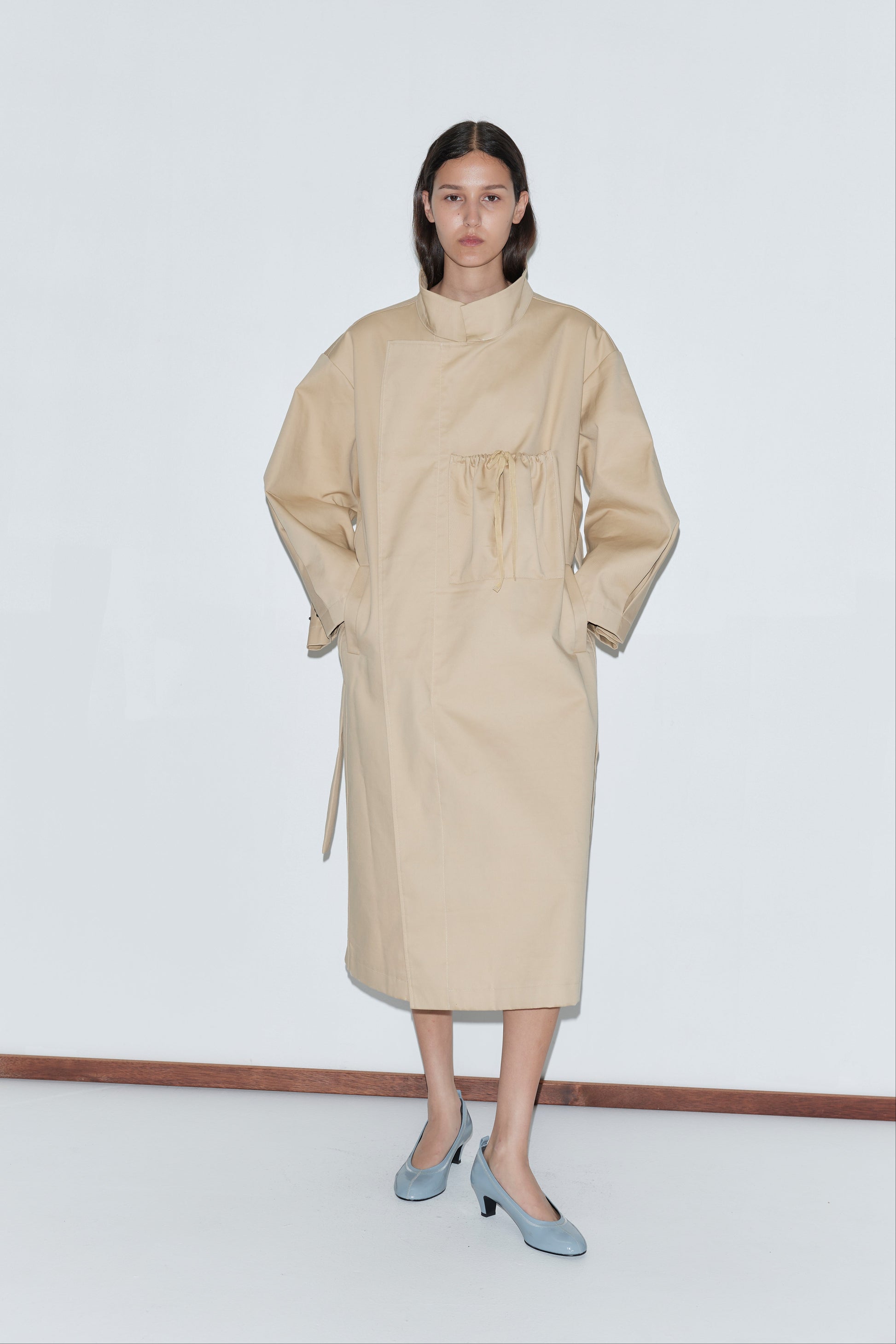 A woman stands against a white wall, wearing the Longline Jacket - Beige with a high collar and gathered waist, paired with light blue heels.