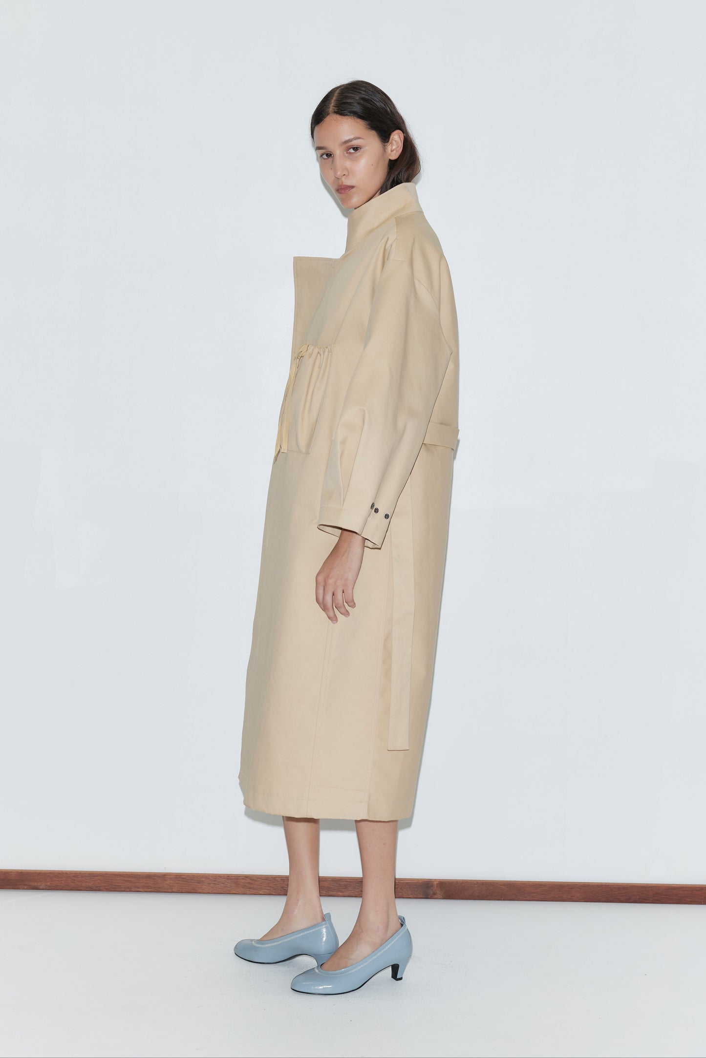 A woman with dark hair stands sideways against a plain white background, wearing the Longline Jacket - Beige and light blue high-heeled shoes.