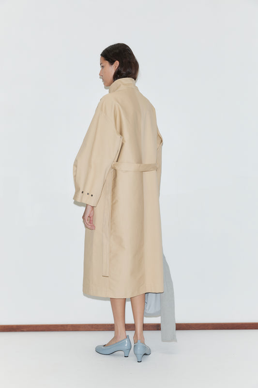 A woman stands facing away, wearing the Longline Jacket - Beige by Deiji Studios with a back belt and light blue heels. She holds a light gray garment in her left hand against a plain white background.
