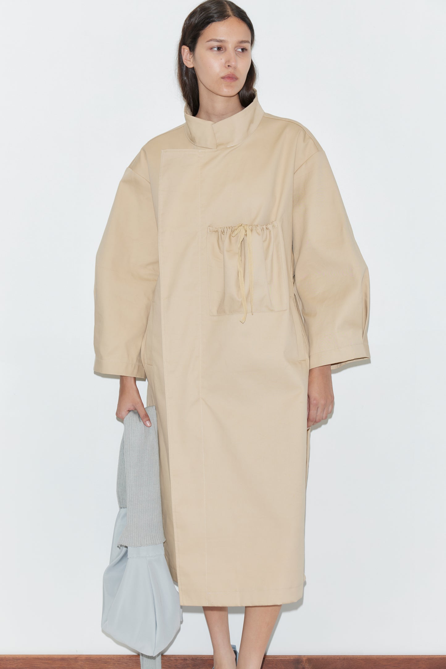 A woman stands against a plain white wall wearing the Longline Jacket - Beige from Deiji Studios, which has a drawstring chest pocket. She holds a light gray bag and a folded light garment in her left hand.