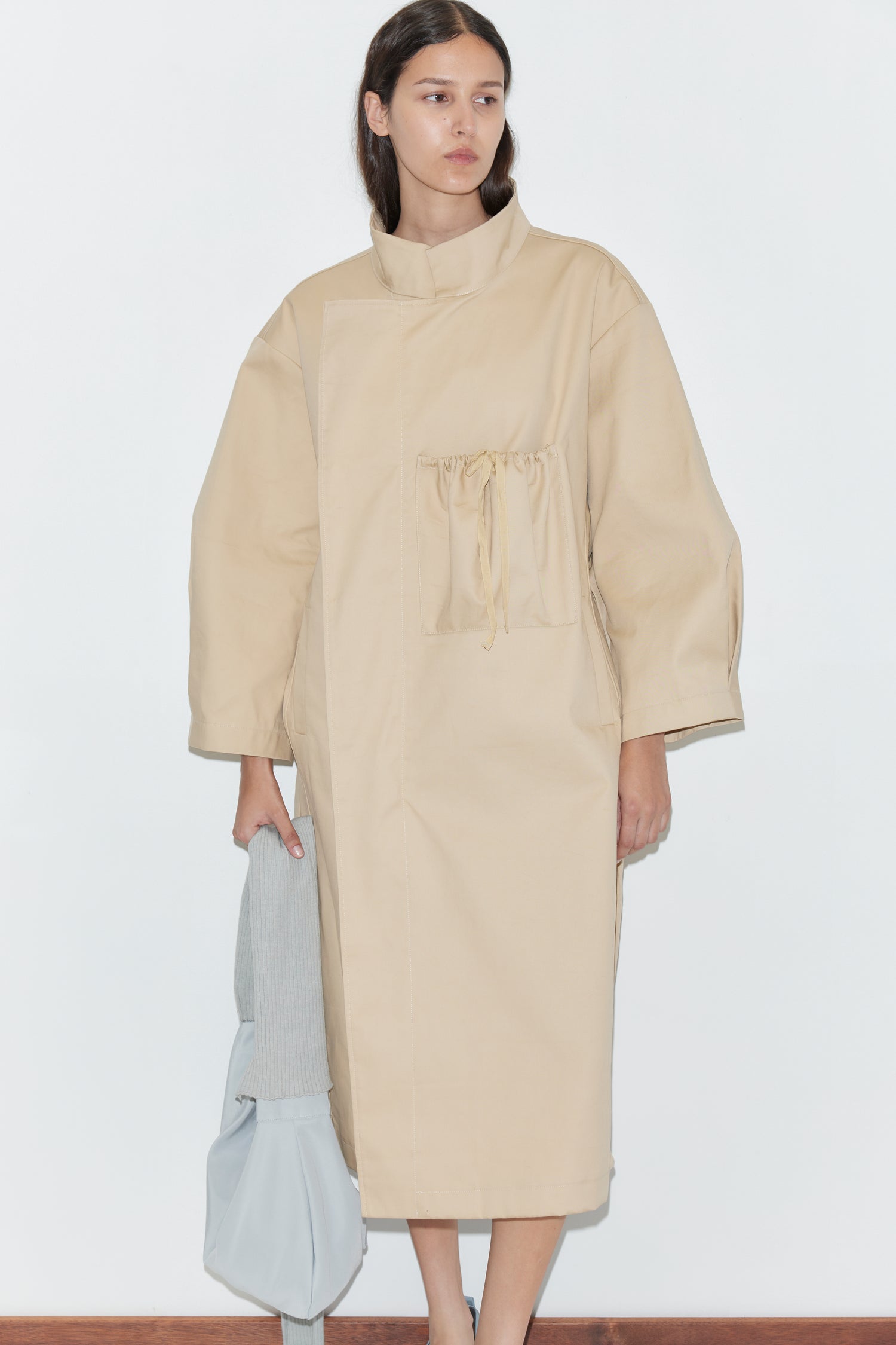 A woman stands against a plain white wall wearing the Longline Jacket - Beige from Deiji Studios, which has a drawstring chest pocket. She holds a light gray bag and a folded light garment in her left hand.