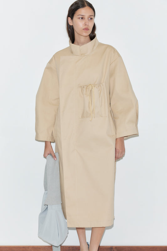 A woman stands against a plain white wall wearing the Longline Jacket - Beige from Deiji Studios, which has a drawstring chest pocket. She holds a light gray bag and a folded light garment in her left hand.
