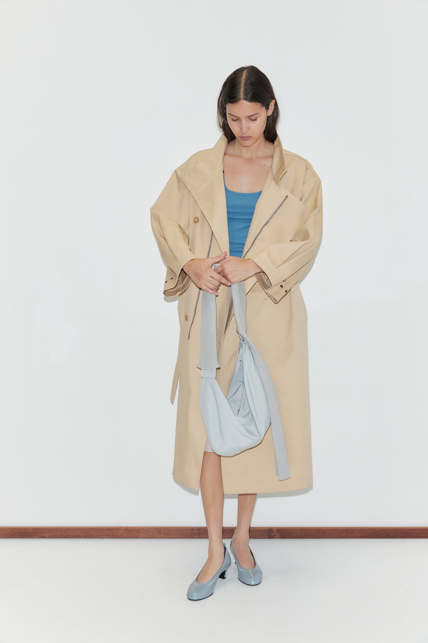 A woman stands against a white background wearing the Longline Jacket - Beige from Deiji Studios, paired with a blue top and blue heels. She looks down while adjusting a light gray sling-style bag on her shoulder.