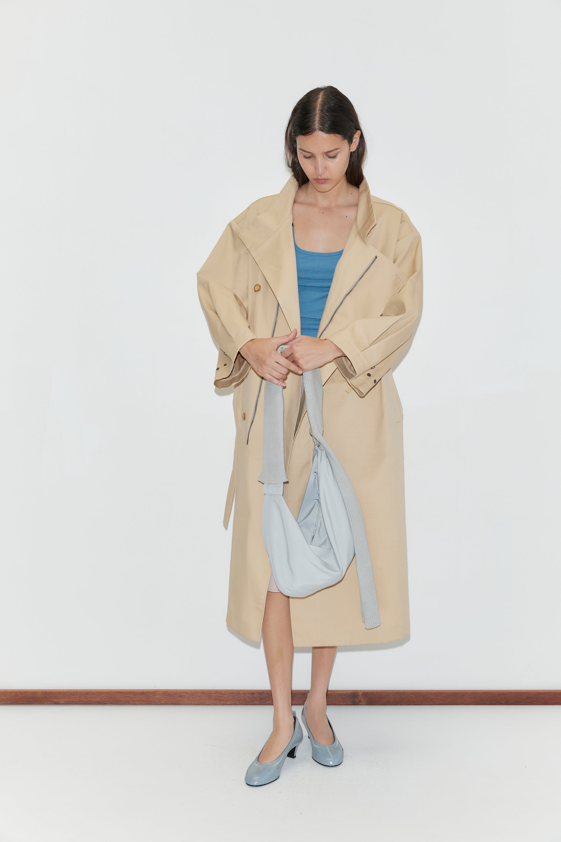 A woman stands against a white background wearing the Longline Jacket - Beige from Deiji Studios, paired with a blue top and blue heels. She looks down while adjusting a light gray sling-style bag on her shoulder.