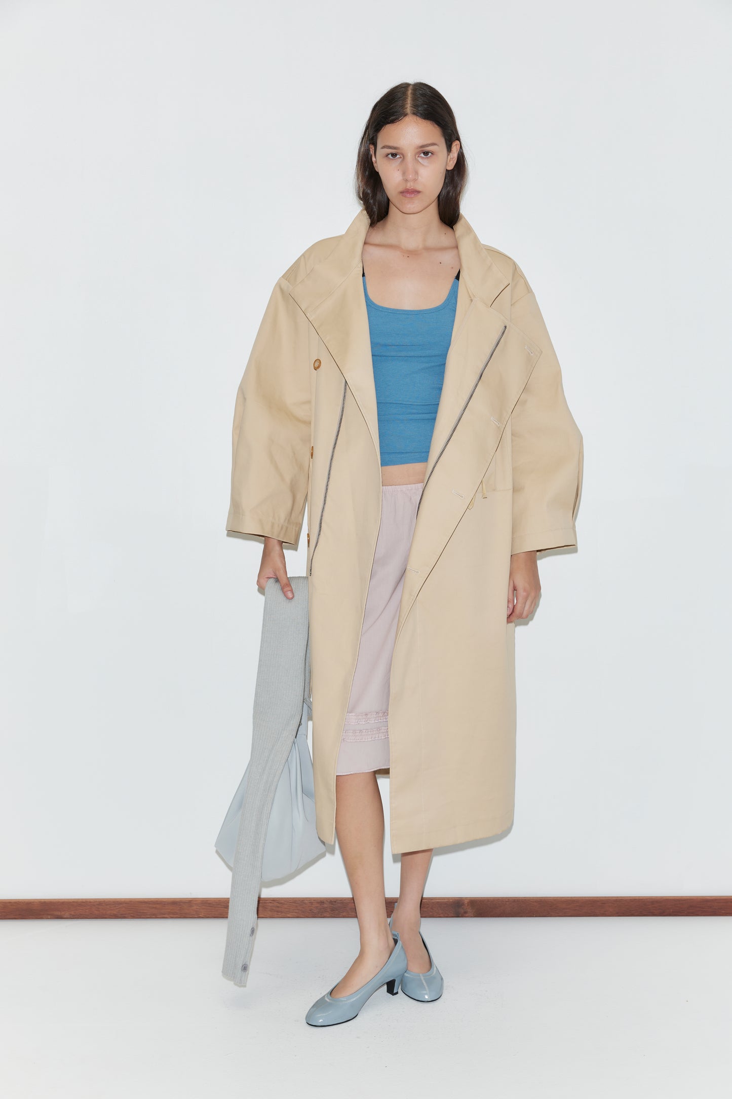 A woman stands against a white wall, wearing a beige trench coat over a blue top and the Daytime Skirt - Floss, a light pink cotton voile skirt. She holds a light gray garment, wears light blue shoes, has loose hair, and shows a neutral expression.