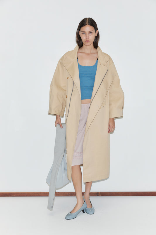 A woman stands against a white wall, wearing a beige trench coat over a blue top and the Daytime Skirt - Floss, a light pink cotton voile skirt. She holds a light gray garment, wears light blue shoes, has loose hair, and shows a neutral expression.