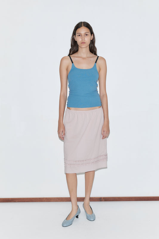 A woman with long dark hair wears a fitted blue camisole, the Daytime Skirt - Floss in pale pink cotton voile, and light blue low-heeled shoes, standing against a plain white background.