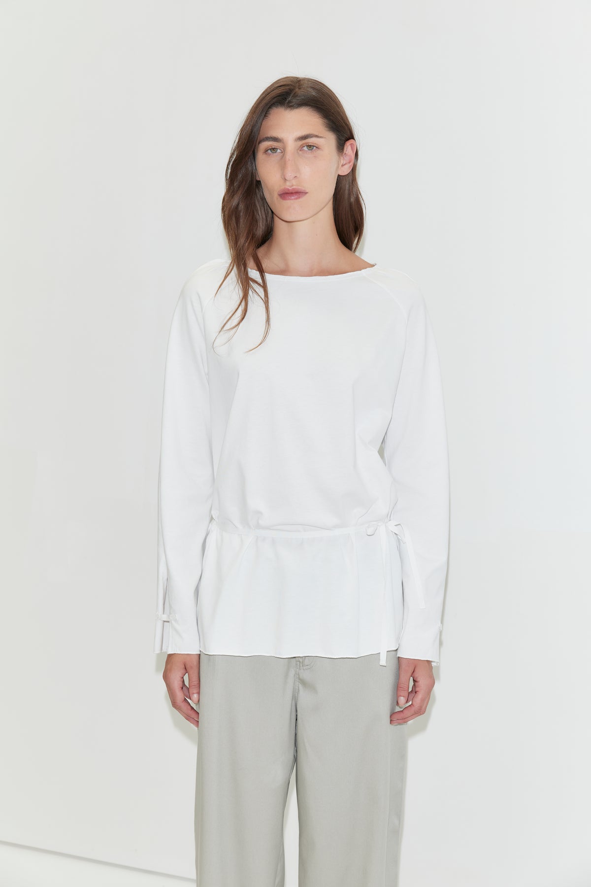 A woman with long dark hair stands against a plain white background wearing the Wided Longsleeve - White by Deiji Studios, a white top with a wide neckline and drawstring hem, with grey wide-leg trousers.