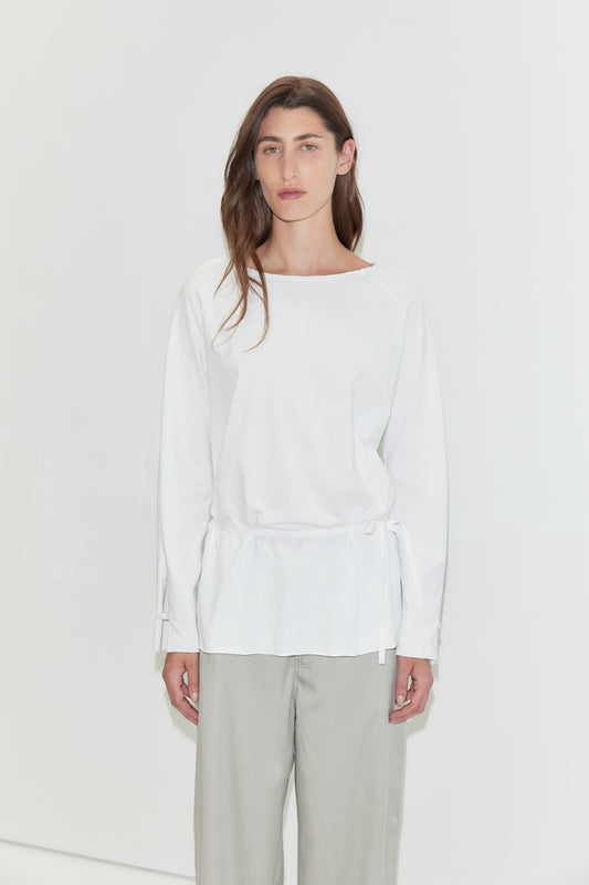 A woman with long dark hair stands against a plain white background wearing the Wided Longsleeve - White by Deiji Studios, a white top with a wide neckline and drawstring hem, with grey wide-leg trousers.