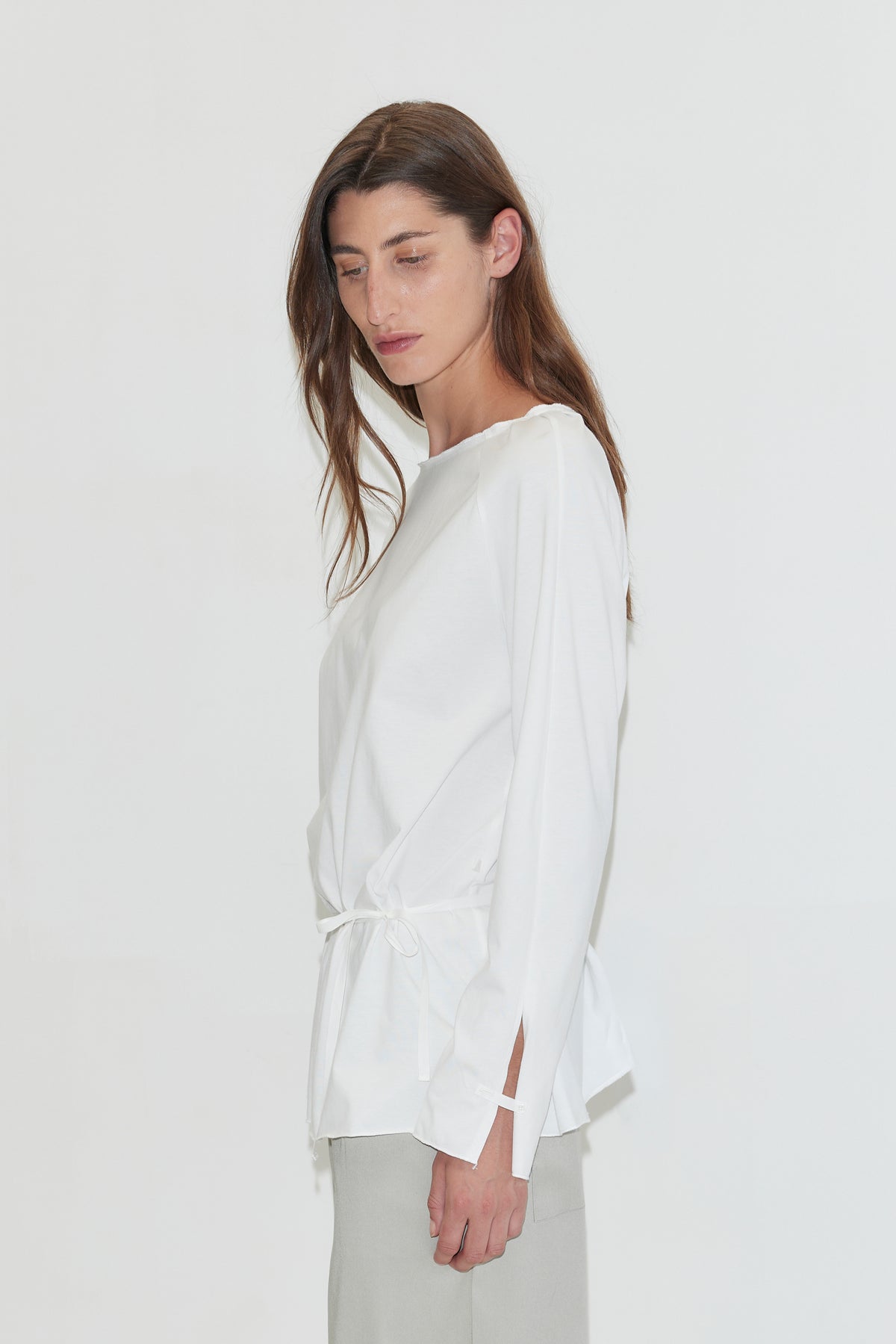 A woman with long dark hair stands in profile against a plain white background, looking down, wearing the Wided Longsleeve - White by Deiji Studios, a white top with a wide neckline and drawstring hem, with grey trousers.
