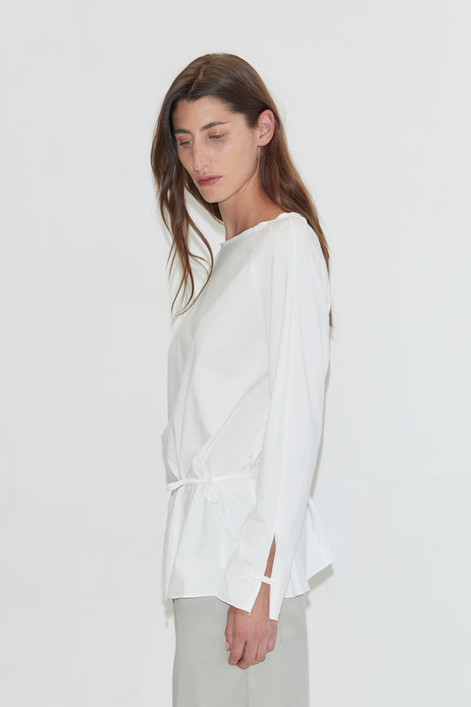 A woman with long dark hair stands in profile against a plain white background, looking down, wearing the Wided Longsleeve - White by Deiji Studios, a white top with a wide neckline and drawstring hem, with grey trousers.