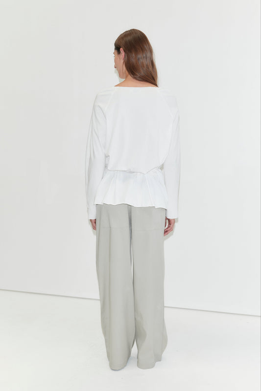 A woman with long dark hair stands with her back to the camera against a plain white background wearing the Wided Longsleeve - White by Deiji Studios, a white top with a gathered hem, with grey wide-leg trousers.