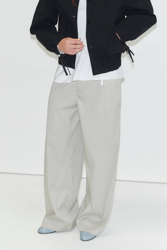 A person wearing a black jacket, white shirt, Tencel Pant - Grey by Deiji Studios, and light blue pointed shoes stands against a plain white background. Their head is not visible in the image.