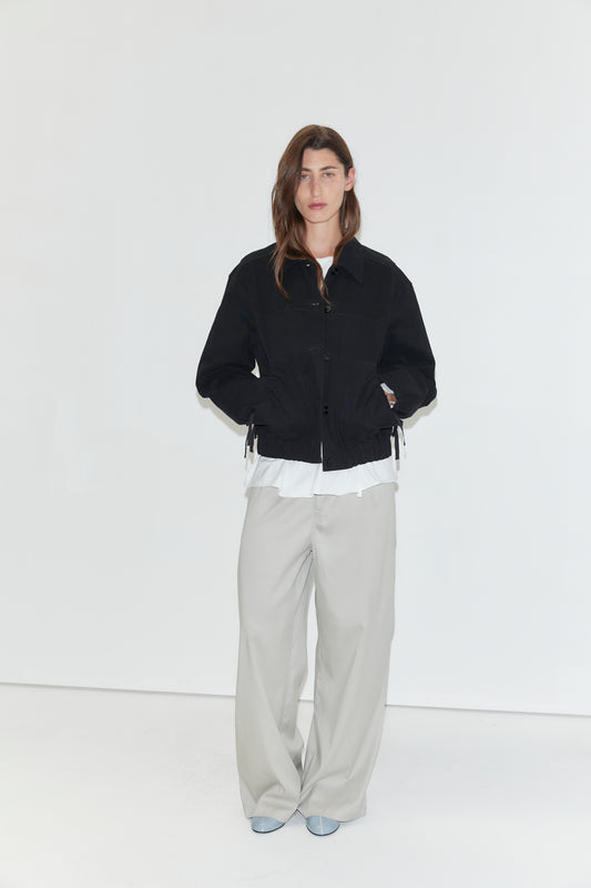 A person stands against a plain white background, wearing a black jacket over a white shirt, the Tencel Pant - Grey from Deiji Studios styled as loose wide-leg pants, and light blue shoes, with their hands in their jacket pockets.