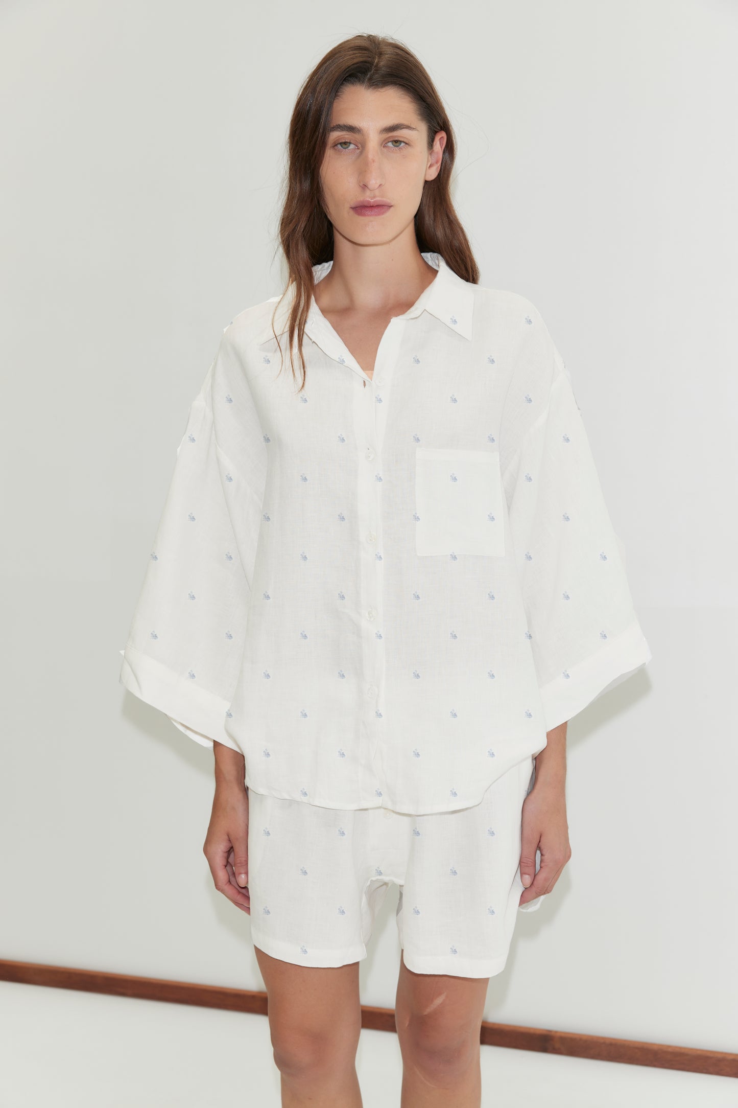 A person with long brown hair stands against a plain white background, wearing Deiji Studios’ 03 Set - Floral Blue: an oversized linen shirt with subtle small patterns and matching boxer-style shorts.