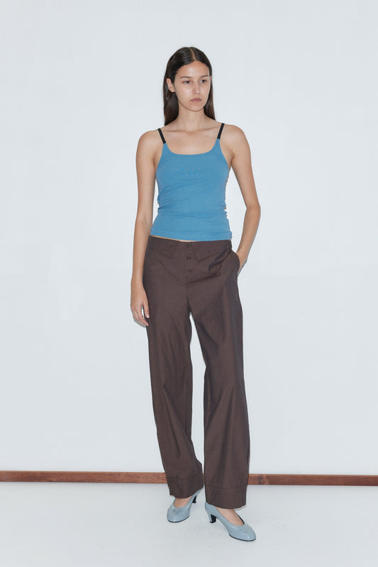 A woman stands against a plain white wall wearing a blue tank top, Collar Point Pant in Toast by Deiji Studios, and light blue heels. One hand is in her pocket and she has a neutral expression.