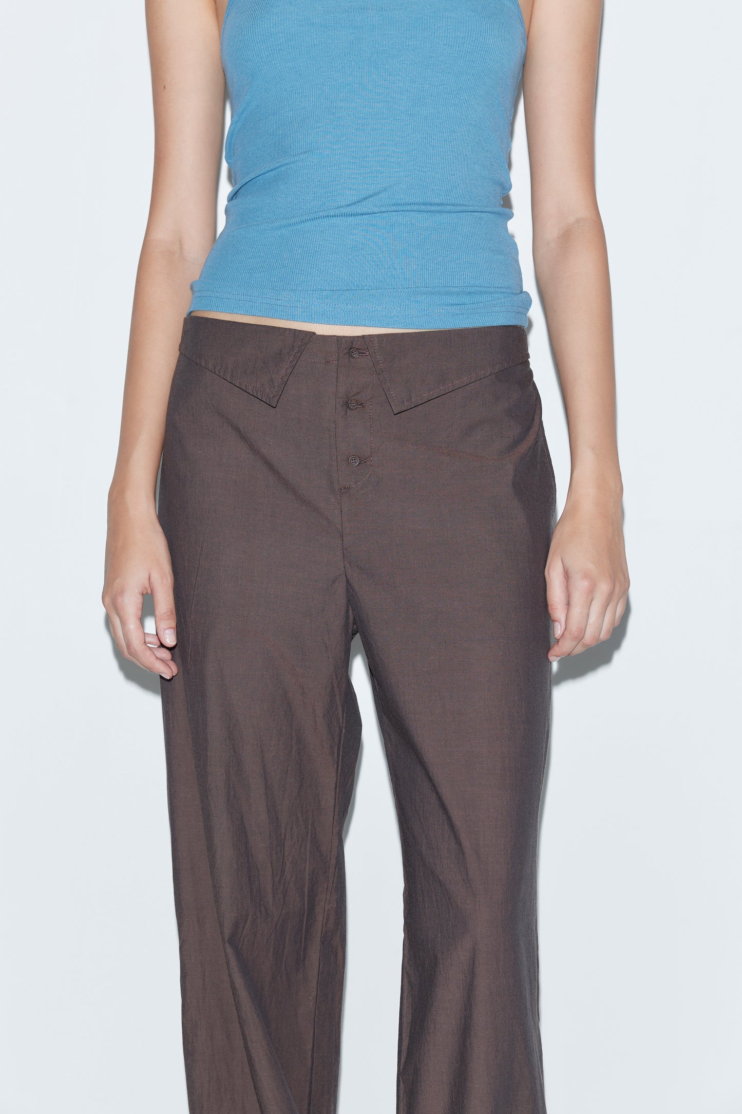 A person wears a light blue sleeveless top and the Collar Point Pant - Toast by Deiji Studios, standing against a plain light background and shown from shoulders to mid-thigh.