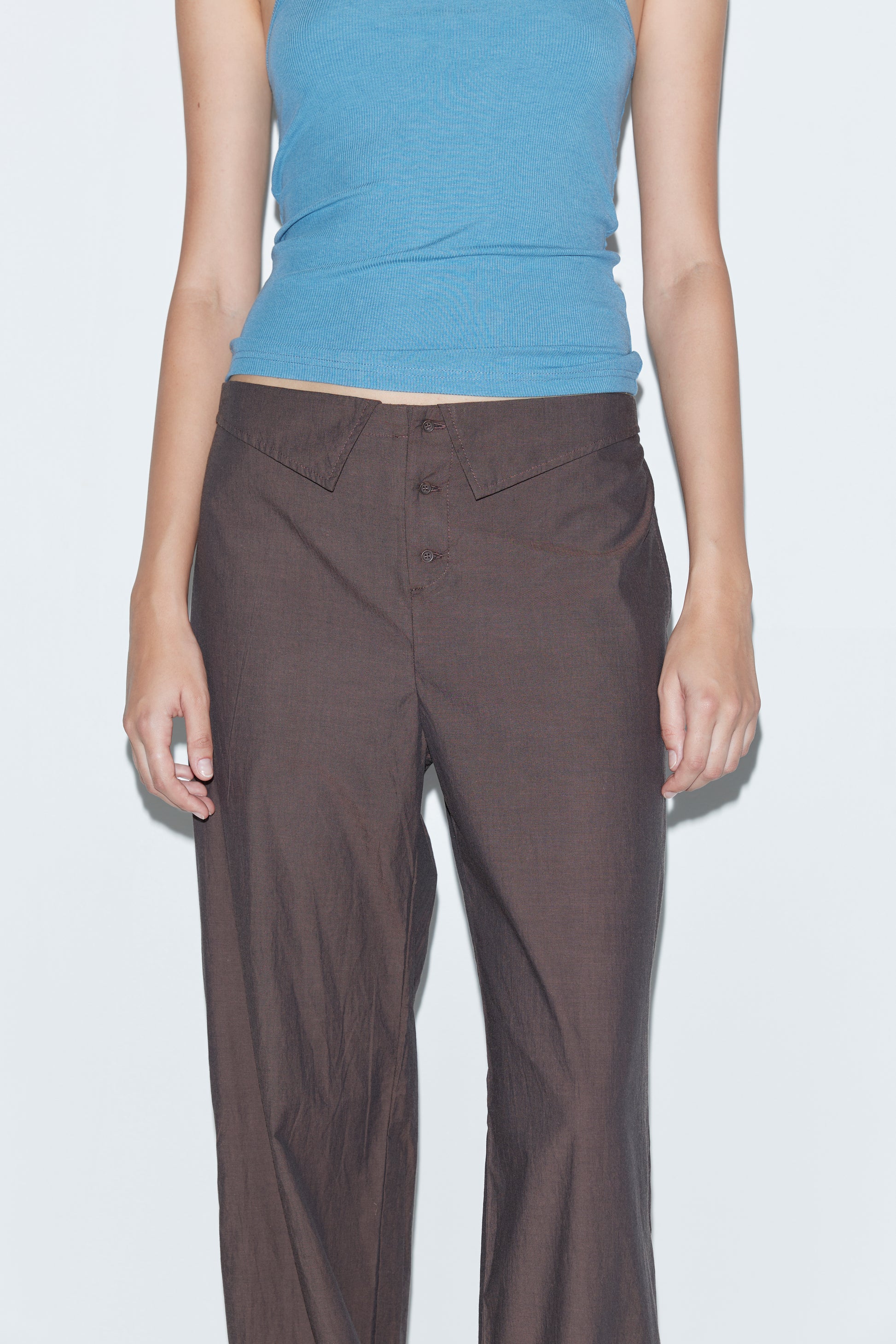 A person wears a light blue sleeveless top and the Collar Point Pant - Toast by Deiji Studios, standing against a plain light background and shown from shoulders to mid-thigh.