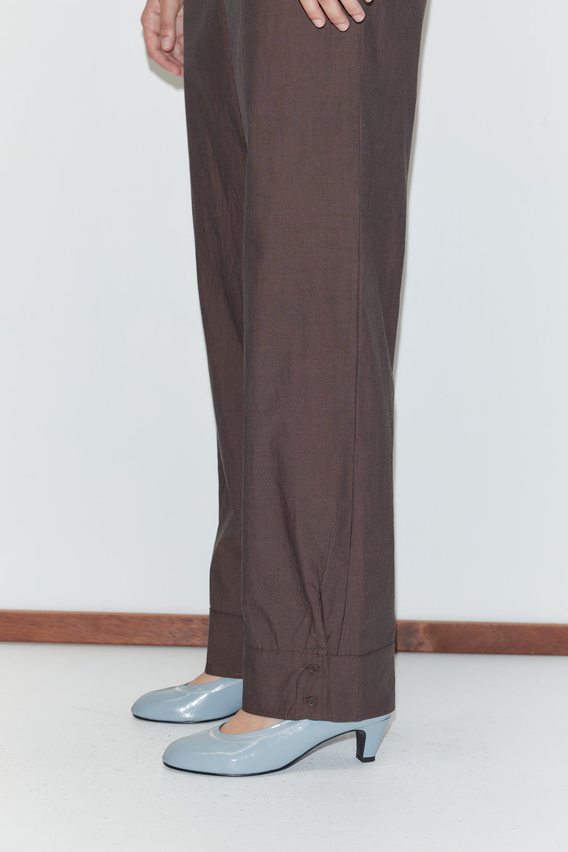 A person wearing Deiji Studios' Collar Point Pant in Toast and light blue, low-heeled shoes stands on a white floor by a white wall with a wooden baseboard, with only their lower body visible.