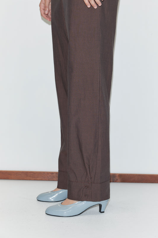 A person wearing Deiji Studios' Collar Point Pant in Toast and light blue, low-heeled shoes stands on a white floor by a white wall with a wooden baseboard, with only their lower body visible.
