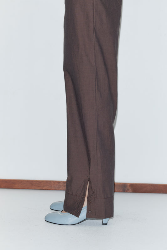 A person wearing Deiji Studios’ Collar Point Pant - Toast with ankle slits and light blue low-heeled shoes stands against a white wall and floor with wooden trim.