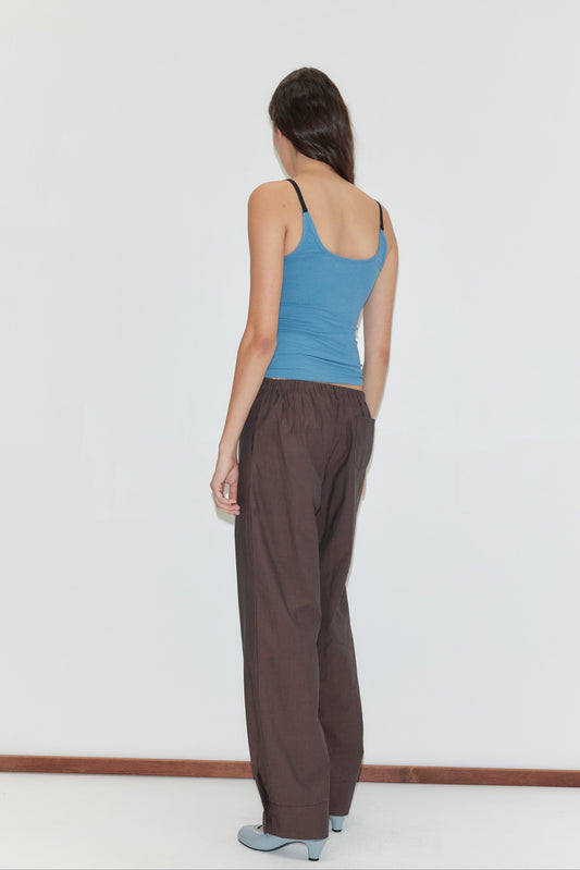 A woman with long brown hair faces away, wearing a blue tank top, Collar Point Pant - Toast from Deiji Studios, and light blue shoes against a plain white background.