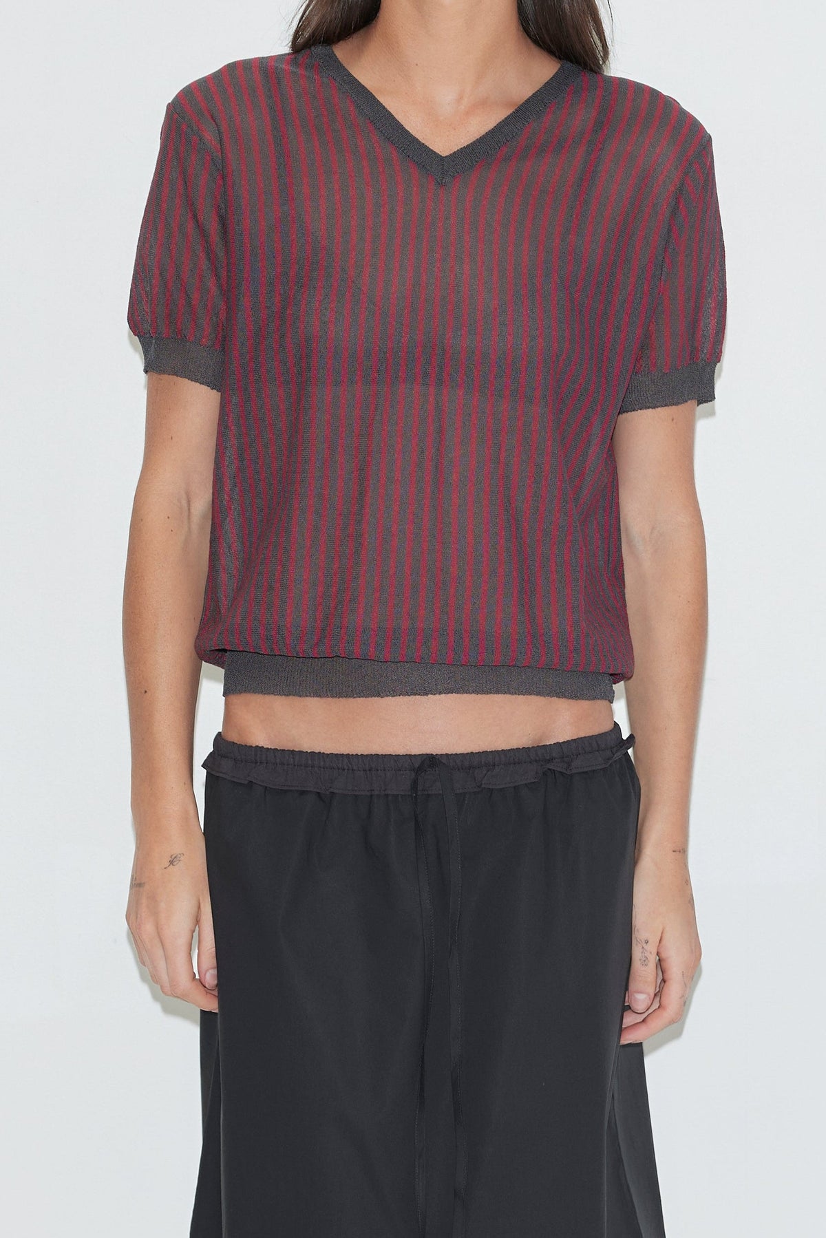 A cropped front view of the Cross Neck Tee - Red Stripe by Deiji Studios, a fine knit tee with red and charcoal vertical stripes, a V-neckline and ribbed trims, worn with a black skirt against a plain white background.