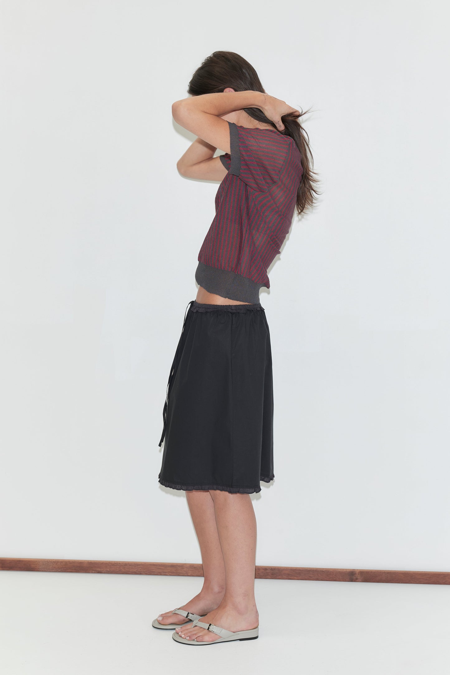 A woman with long dark hair stands in profile against a plain white background wearing the One Frill Skirt - Black by Deiji Studios, a black knee-length skirt with a drawcord waist and frill hem, with a red striped knit tee and flip-flops.