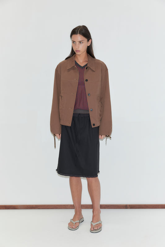 A woman with long dark hair stands against a plain white background wearing the One Frill Skirt - Black by Deiji Studios, a black knee-length skirt with a frill hem, with a red striped knit tee, a brown jacket and flip-flops.