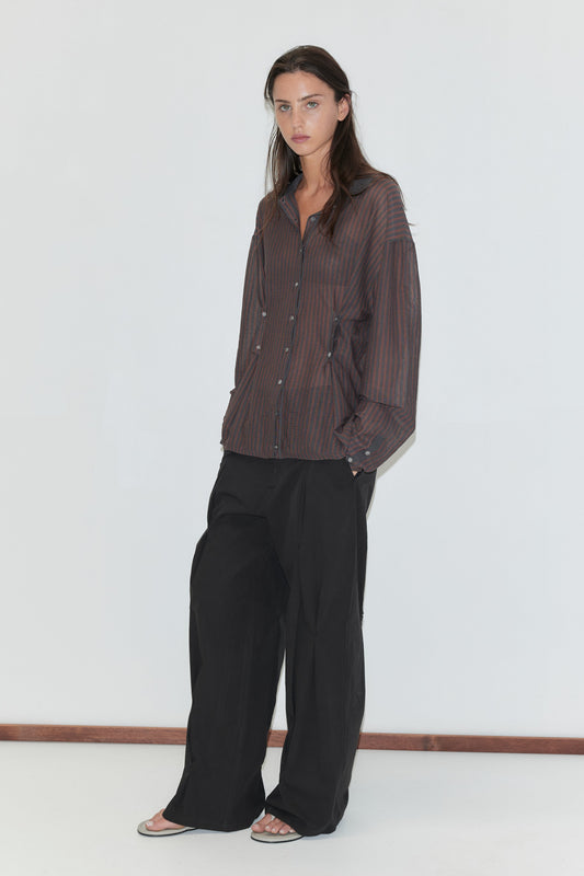 A woman with long dark hair stands against a plain white background wearing the Multi Pleat Pant - Black by Deiji Studios, black wide-leg pleated trousers, with a sheer brown striped knit shirt and flip-flops.