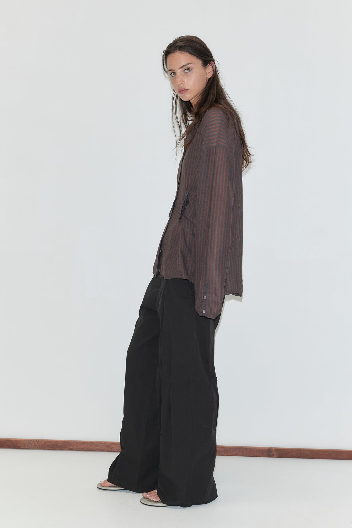 A woman with long dark hair stands in profile against a plain white background wearing the Multi Pleat Pant - Black by Deiji Studios, black wide-leg pleated trousers, with a sheer brown striped knit shirt.
