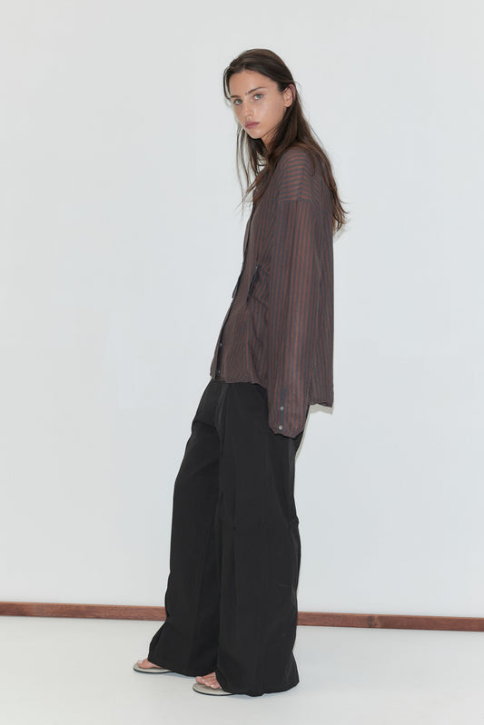 A woman with long dark hair stands in profile against a plain white background wearing the Multi Pleat Pant - Black by Deiji Studios, black wide-leg pleated trousers, with a sheer brown striped knit shirt.