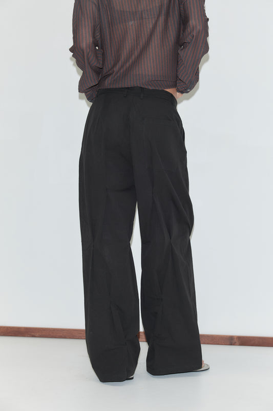 A cropped back view of the Multi Pleat Pant - Black by Deiji Studios, black wide-leg pleated trousers with back welt pockets, worn with a sheer brown striped knit shirt against a plain white background.