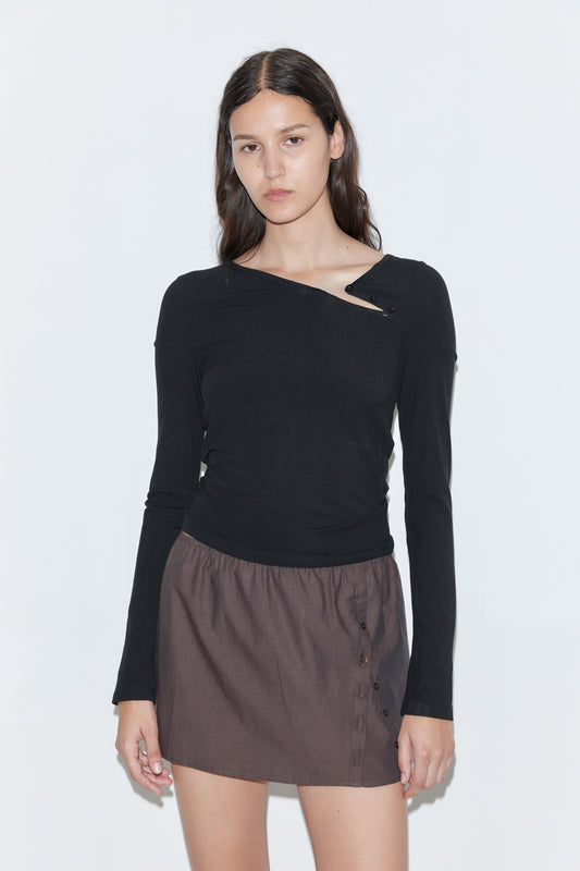 A young woman with long dark hair wears a black long-sleeve top and the Pleat Split Skirt in Toast, standing against a plain white background and looking at the camera with a neutral expression.