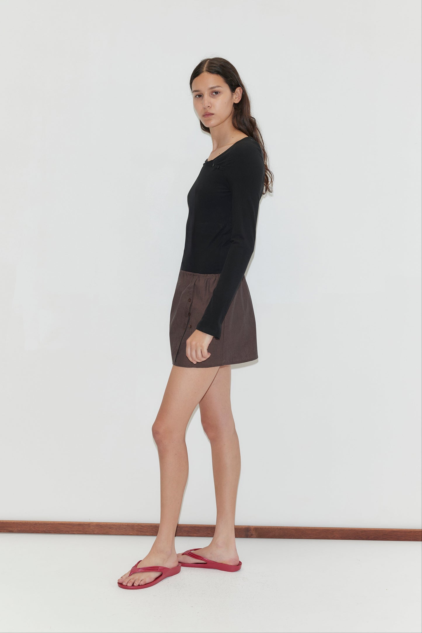 A woman with long dark hair stands sideways against a plain white background, wearing the Multi Button Top - Black with long sleeves, paired with brown shorts and red flip-flops.
