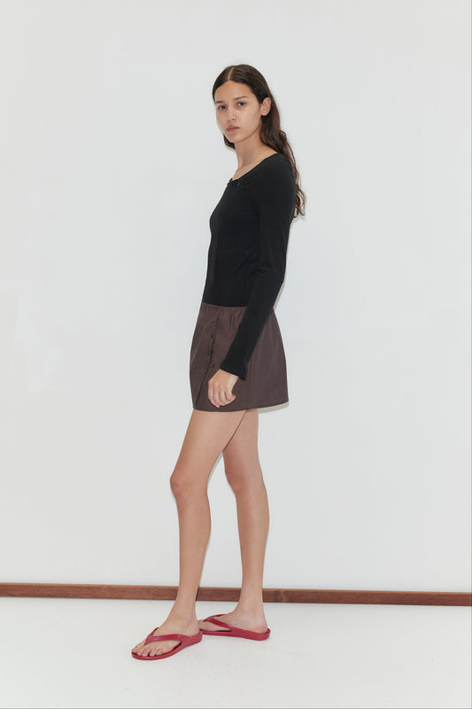 A woman with long dark hair stands sideways against a plain white background, wearing the Multi Button Top - Black with long sleeves, paired with brown shorts and red flip-flops.