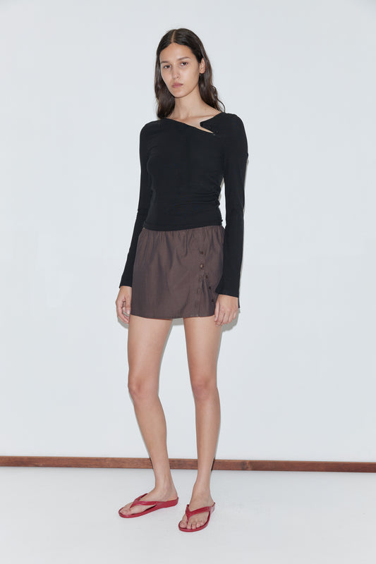 A woman with long brown hair and a neutral expression stands against a white wall, wearing a black off-shoulder long-sleeve top, the Pleat Split Skirt - Toast with an elasticated waistband, and red flip-flops.